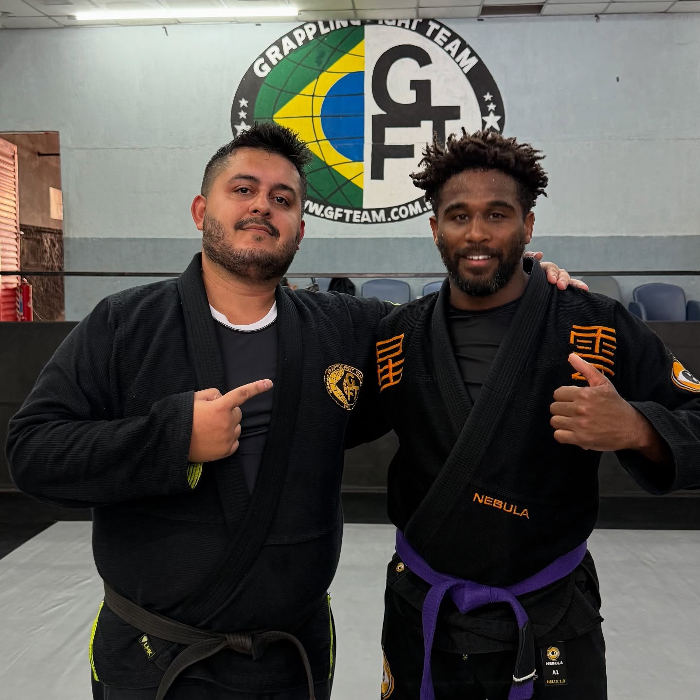 Two black belts in gis in front of the GFTeam logo wall in Rio