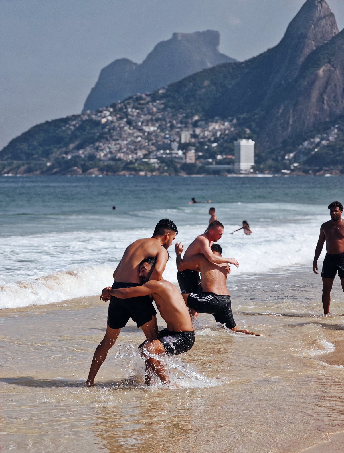 OSS Rio Tour travelers grappling in the surf on a Rio de Janeiro beach, with Dois Irmãos and the Vidigal hillside behind them